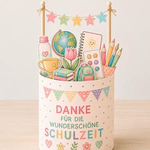 May include: A cylindrical container with a cream background and pastel bunting, featuring the text "DANKE FÜR DIE WUNDERSCHÖNE SCHULZEIT". It holds cutouts of a globe, trophy, books, backpack, paint palette, pencils, and a tulip, topped with a star banner.