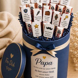 May include: A navy blue gift box with a gold ribbon and bow, filled with chocolate bars and cards. The cards have various messages and icons, including a trophy, car, and heart. The box lid says "F&uuml;r Dich" and the box says "Papa".