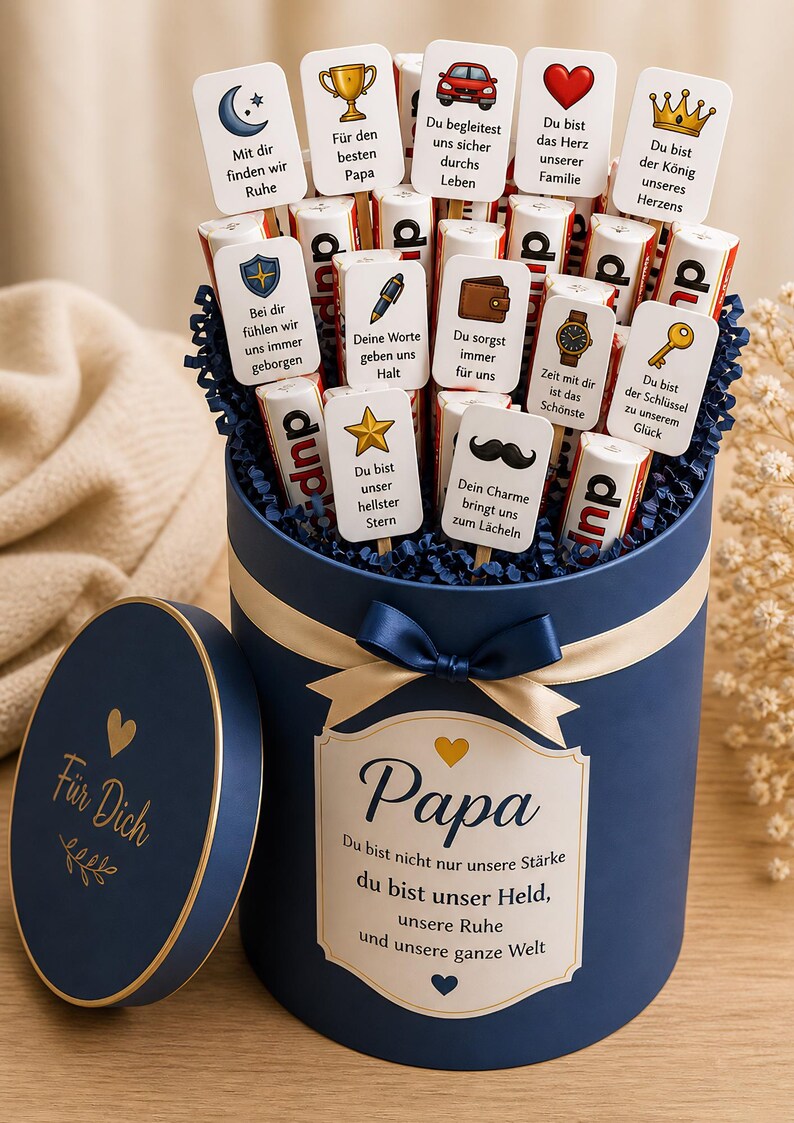 May include: A navy blue gift box with a gold ribbon and bow, filled with chocolate bars and cards. The cards have various messages and icons, including a trophy, car, and heart. The box lid says "F&uuml;r Dich" and the box says "Papa".