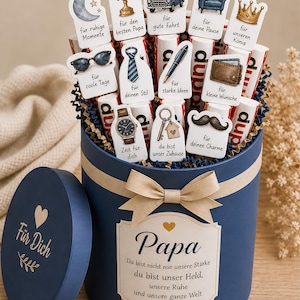 Puede incluir: Una caja de regalo azul marino con tapa, llena de barras de chocolate y recortes decorativos. Los recortes presentan varias imágenes y frases, incluyendo un coche, un reloj y la palabra "Papa". Un lazo adorna la parte delantera de la caja.