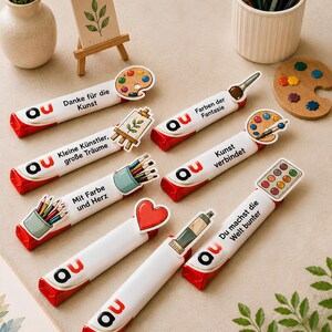 May include: Seven rectangular candy bars wrapped in white paper with red ends, each featuring the logo 'au' and various artistic illustrations including paint palettes, brushes, pencils, and hearts, with German text printed on each individual wrapper.