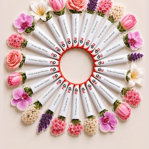 Op de afbeelding: Een cirkelvormige opstelling van vierentwintig rechthoekige witte chocoladerepen, elk verpakt in papier met een unieke Duitse zin en bekroond met een andere kleurrijke bloem, waaronder roze rozen, paarse lavendel en witte lelies.