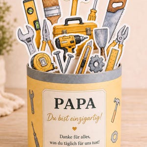 以下が含まれることがあります： 「PAPA」と「Du bist einzigartig!」と書かれたラベルが付いた黄色の容器で、さまざまな工具の切り抜きがあります。 工具には、ハンマー、のこぎり、水準器、ドライバーが含まれます。 テキスト「Danke für alles, was du täglich für uns tust!」も存在します。