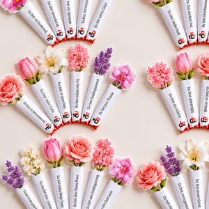 Op de afbeelding: Een verzameling rechthoekige chocoladerepen verpakt in wit papier, elk bekroond met een ander driedimensionaal bloemstuk met rozen, tulpen, lelies en lavendel. Elke wikkel is voorzien van een Duitse tekst en een klein rood logo.