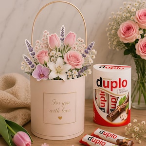 Pode incluir: Uma caixa de presente cilíndrica rosa-claro com uma alça dourada, contendo um arranjo floral de rosas cor-de-rosa, tulipas e lavanda. A caixa apresenta o texto "For you with love" e um ícone de coração. Ao lado, há uma embalagem de Ferrero duplo.