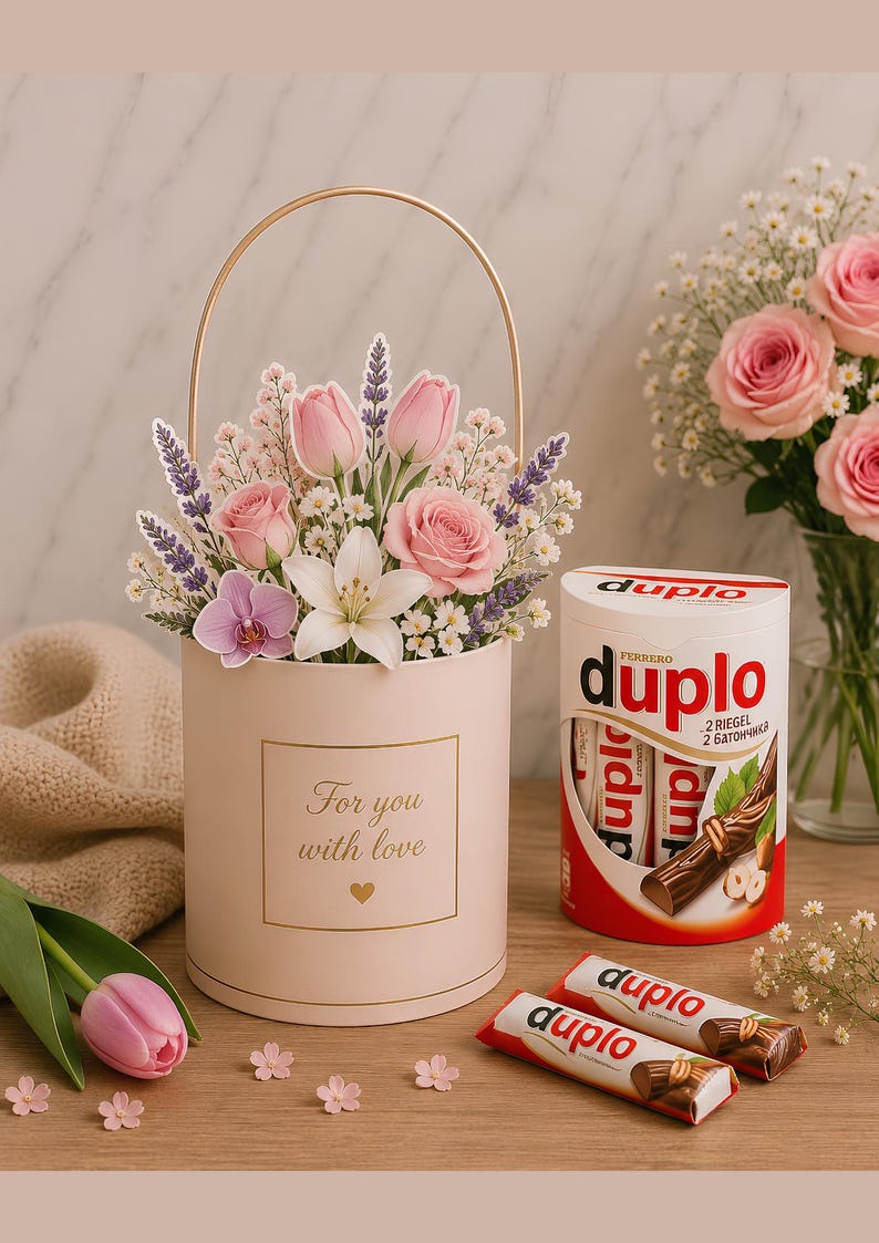 Op de afbeelding: Een cilindervormige lichtroze geschenkdoos met een gouden handvat, gevuld met een bloemstuk van roze rozen, tulpen en lavendel. De doos bevat de tekst "For you with love" en een hartje. Daarnaast staat een verpakking met Ferrero duplo chocolade.