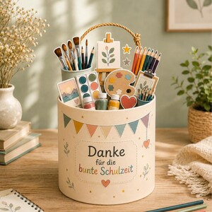 May include: A cylindrical cream-colored gift box with a rope handle, decorated with colorful bunting and the text "Danke f&uuml;r die bunte Schulzeit". The box is filled with art supplies including paintbrushes, colored pencils, paints, and small art-themed cutouts.