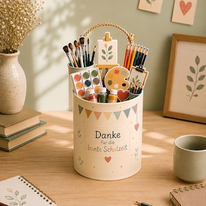 Peut inclure: Un récipient cylindrique de couleur crème avec une poignée en corde, décoré de fanions colorés et du texte "Danke für die bunte Schulzeit". Il contient diverses fournitures d'art, dont des pinceaux, des crayons de couleur, des pots de peinture.