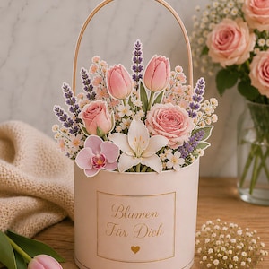 Op de afbeelding: Een cilindervormige lichtroze geschenkdoos met een gouden handvat en de tekst Blumen F&uuml;r Dich op de voorzijde. De doos bevat een papieren uitgesneden arrangement van roze tulpen, rozen, lavendeltakjes, witte lelies en kleine witte bloemen.