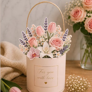 Op de afbeelding: Een cilindervormige lichtroze geschenkdoos met een gouden handvat, voorzien van een bloemstuk met roze rozen, tulpen, een witte lelie, een orchidee en paarse lavendel. De doos heeft een goudomrand label met de tekst For you with love en een hartje.