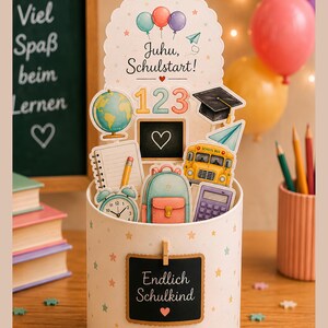 May include: A cylindrical gift container decorated with small stars, featuring a cutout topper with balloons and the text Juhu, Schulstart! It includes illustrations of a globe, numbers 123, a graduation cap, a school bus, a backpack, and a clock. A small chalkboard sign on the front reads Endlich Schulkind.