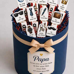 May include: A navy blue gift box with a gold ribbon and bow, filled with chocolate bars and decorative toppers. The toppers feature various illustrations and phrases, including a trophy, car, and the word "Papa".