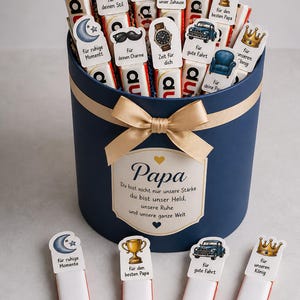 May include: A navy blue gift box with a gold bow, filled with chocolate bars and decorative toppers. The toppers feature various designs and phrases, including a tie, mustache, and car. The box has a label that says "Papa".