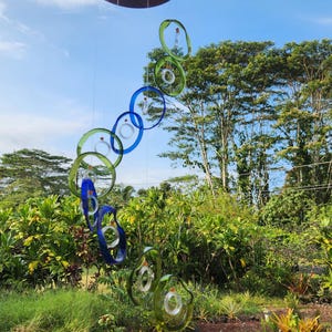 May include: A colorful outdoor wind chime with a series of glass rings in shades of blue and green, suspended from a brown disc. The wind chime hangs in front of lush green foliage and a blue sky.
