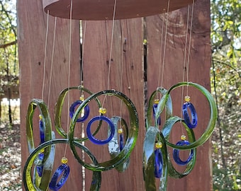 May include: A green glass wind chime with blue glass rings and yellow beads hanging from a wooden oval frame.