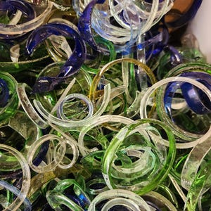 May include: A close-up shot of a collection of glass rings in various colors. The rings are clear, blue, green, and yellow. The glass rings are in different sizes and shapes, some are curved, and some are circular.