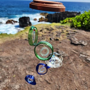 May include: A wind chime made of recycled glass bottles. The chime has a wooden top and hangs from a string. The glass bottles are green and blue and are shaped into circles and cones. The chime is hanging in front of a blue ocean and green grass.