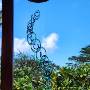 May include: A hanging wind chime with a dark brown circular top and a series of clear and blue glass rings suspended below. The wind chime is hanging in front of a blue sky and lush green foliage.