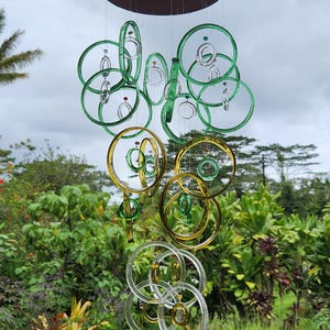 May include: A hanging wind chime with a dark brown circular top and multiple clear, green, and yellow glass rings. The rings are suspended by thin wires, creating a decorative outdoor ornament. The background features lush green foliage and a cloudy sky.