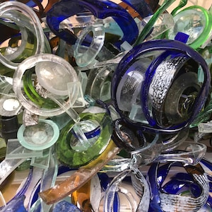 May include: A close-up of a pile of recycled glass bottle pieces. The pieces are in various shapes and sizes, including circles, squares, and rectangles. The colors of the glass are clear, green, and blue.