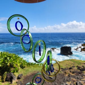 May include: A handcrafted wind chime featuring green and blue glass rings suspended from a wooden disc. The rings vary in size and are connected with thin strings. The background shows a blue ocean and sky.