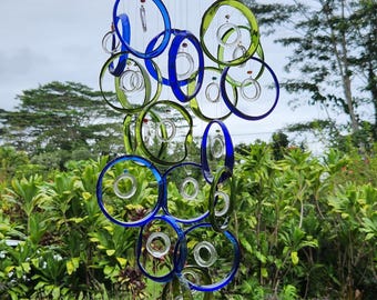 May include: A handcrafted wind chime featuring a series of blue and green glass circles suspended from a brown wooden disc. The circles are connected by clear strings, creating a decorative outdoor accent. The background shows lush green foliage.
