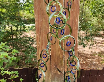 May include: A handcrafted wind chime featuring green glass rings with blue glass accents, suspended from a wooden base. The rings are connected by clear strings, creating a cascading effect. The wind chime is displayed outdoors, with a natural background.