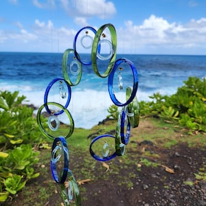 May include: A handcrafted wind chime featuring a series of oval-shaped glass rings in shades of blue and green. Each ring is suspended by a thin string, with small glass accents. The background shows a blue ocean and sky.