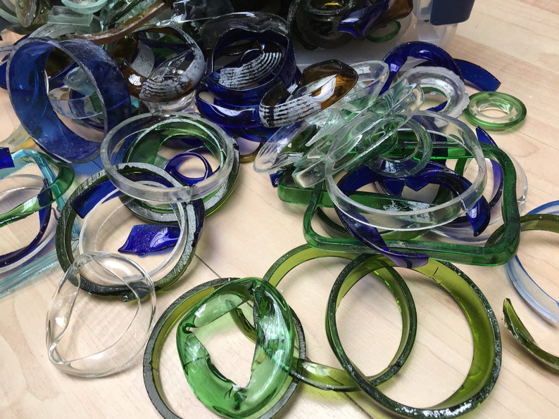 May include: A pile of glass bottle rings in various shades of blue, green, and clear. The rings are arranged in a random pattern on a wooden surface.