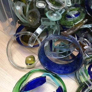 May include: A close-up of a pile of broken glass bottles, with various colors including blue, green, and clear. The glass pieces are in different shapes and sizes, some are circular, some are rectangular, and some are long and thin.