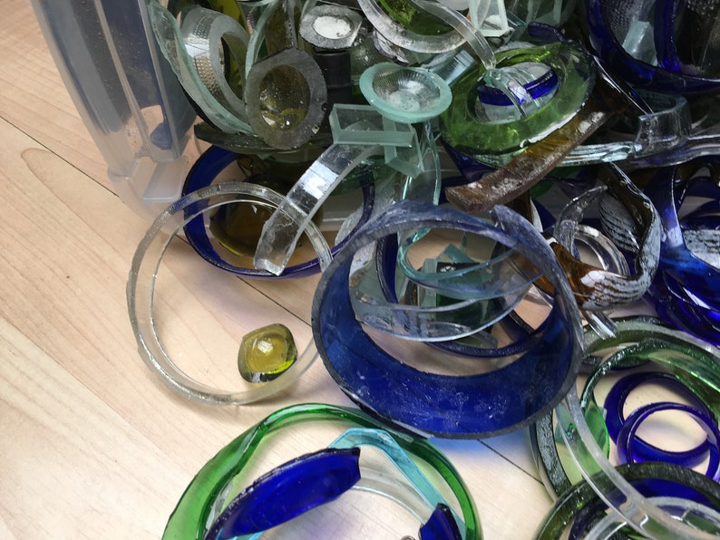 May include: A close-up of a pile of broken glass bottles, with various colors including blue, green, and clear. The glass pieces are in different shapes and sizes, some are circular, some are rectangular, and some are long and thin.