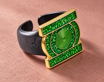 May include: A ring featuring a matte black band with a textured surface and a gold-toned rectangular emblem. The emblem contains a bright green, textured inlay surrounding a central green circular spiral design, creating a bold, geometric look.