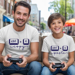 May include: Two white t-shirts with navy blue graphics. One shirt says "ctrl + c copy" and the other says "ctrl + v paste". Both shirts are worn by people holding black game controllers. The shirts are a matching set.