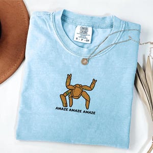May include: A light blue short-sleeve t-shirt with a crew neckline and a Comfort Colors brand tag. The shirt features an embroidered graphic of a brown, multi-legged creature making peace signs with its hands and the text AMAZE AMAZE AMAZE.