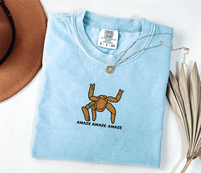 May include: A light blue short-sleeve t-shirt with a crew neckline and a Comfort Colors brand tag. The shirt features an embroidered graphic of a brown, multi-legged creature making peace signs with its hands and the text AMAZE AMAZE AMAZE.
