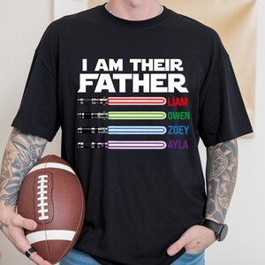 May include: Black t-shirt with the text "I AM THEIR FATHER" in white. Below the text are four lightsabers with the names Liam, Owen, Zoey, and Ayla. A brown football is held in the person's hands.