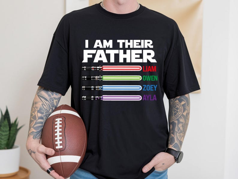 May include: Black t-shirt with the text "I AM THEIR FATHER" in white. Below the text are four lightsabers with the names Liam, Owen, Zoey, and Ayla. A brown football is held in the person's hands.