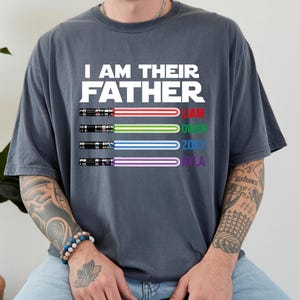 May include: Dark gray t-shirt with the text "I AM THEIR FATHER" in white. Below the text are four lightsabers with the names Liam, Owen, Zoey, and Ayla in red, green, blue, and purple. The shirt is a casual style.