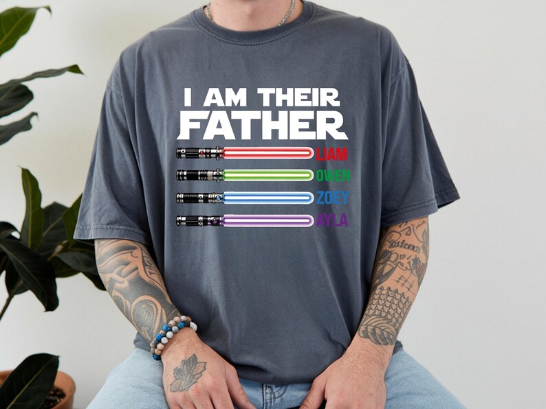 May include: Dark gray t-shirt with the text "I AM THEIR FATHER" in white. Below the text are four lightsabers with the names Liam, Owen, Zoey, and Ayla in red, green, blue, and purple. The shirt is a casual style.