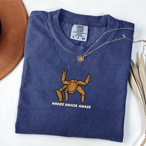 May include: A navy blue short-sleeve t-shirt featuring a graphic of a tan, multi-legged creature with green accents. Below the creature, the text reads AMAZE AMAZE AMAZE in yellow. The shirt has a Comfort Colors brand label at the inner neckline.