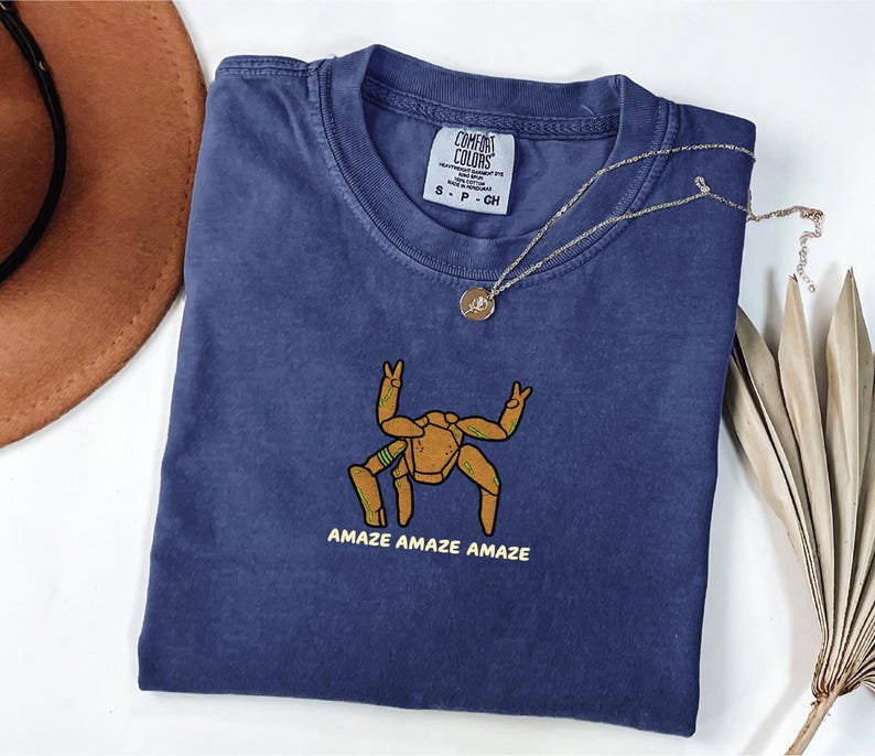 May include: A navy blue short-sleeve t-shirt featuring a graphic of a tan, multi-legged creature with green accents. Below the creature, the text reads AMAZE AMAZE AMAZE in yellow. The shirt has a Comfort Colors brand label at the inner neckline.