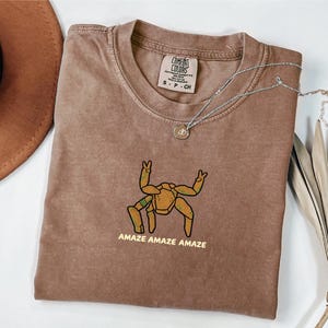 May include: A brown short-sleeve t-shirt featuring a graphic of a tan, multi-legged creature with green accents making peace signs with its upper limbs. The text "AMAZE AMAZE AMAZE" is printed in yellow below the graphic. The shirt has a crew neckline.