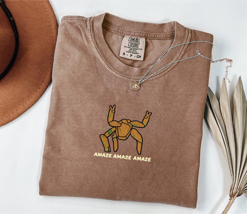 May include: A brown short-sleeve t-shirt featuring a graphic of a tan, multi-legged creature with green accents making peace signs with its upper limbs. The text "AMAZE AMAZE AMAZE" is printed in yellow below the graphic. The shirt has a crew neckline.