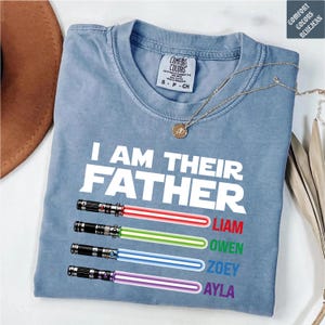 May include: A light blue Comfort Colors t-shirt with the text "I AM THEIR FATHER" and four colored lightsabers with names below. The names are Liam, Owen, Zoey, and Ayla. A gold necklace is draped over the shirt.