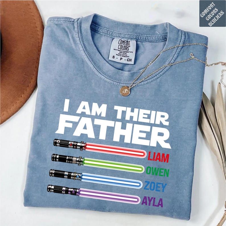 May include: A light blue Comfort Colors t-shirt with the text "I AM THEIR FATHER" and four colored lightsabers with names below. The names are Liam, Owen, Zoey, and Ayla. A gold necklace is draped over the shirt.