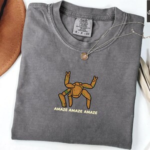 May include: A charcoal gray crew neck t-shirt featuring an embroidered graphic of a tan, multi-legged creature with green accents. Below the creature, the text reads AMAZE AMAZE AMAZE in yellow. The shirt has a Comfort Colors brand label at the neck.