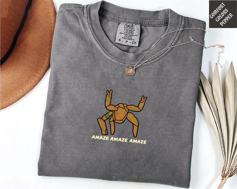 May include: A charcoal gray crew neck t-shirt featuring an embroidered graphic of a tan, multi-legged creature with green accents. Below the creature, the text reads AMAZE AMAZE AMAZE in yellow. The shirt has a Comfort Colors brand label at the neck.