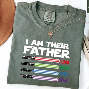 May include: Olive green t-shirt with the text "I AM THEIR FATHER" in white. Below the text are four colorful lightsaber designs with the names Liam, Owen, Zoey, and Ayla in red, green, blue, and purple.