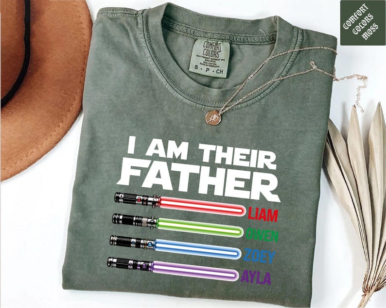 May include: Olive green t-shirt with the text "I AM THEIR FATHER" in white. Below the text are four colorful lightsaber designs with the names Liam, Owen, Zoey, and Ayla in red, green, blue, and purple.