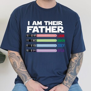May include: Navy blue t-shirt with the text "I AM THEIR FATHER" in white. Below the text are four lightsabers with the names Liam, Owen, Zoey, and Ayla. The lightsabers have red, green, blue, and purple blades.
